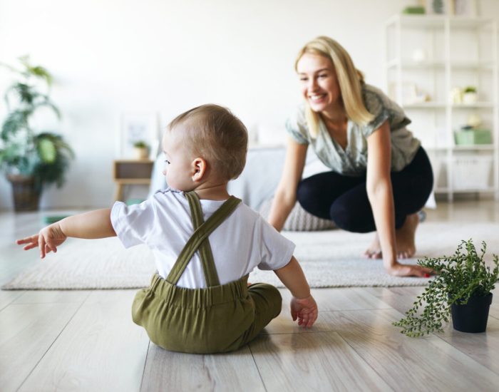 Back view of infant child in stylish clothes sitting on floor with plant pot, raising hand, pointing finger sideways, showing something to his young cheerful mother. Family, motherhood and infancy